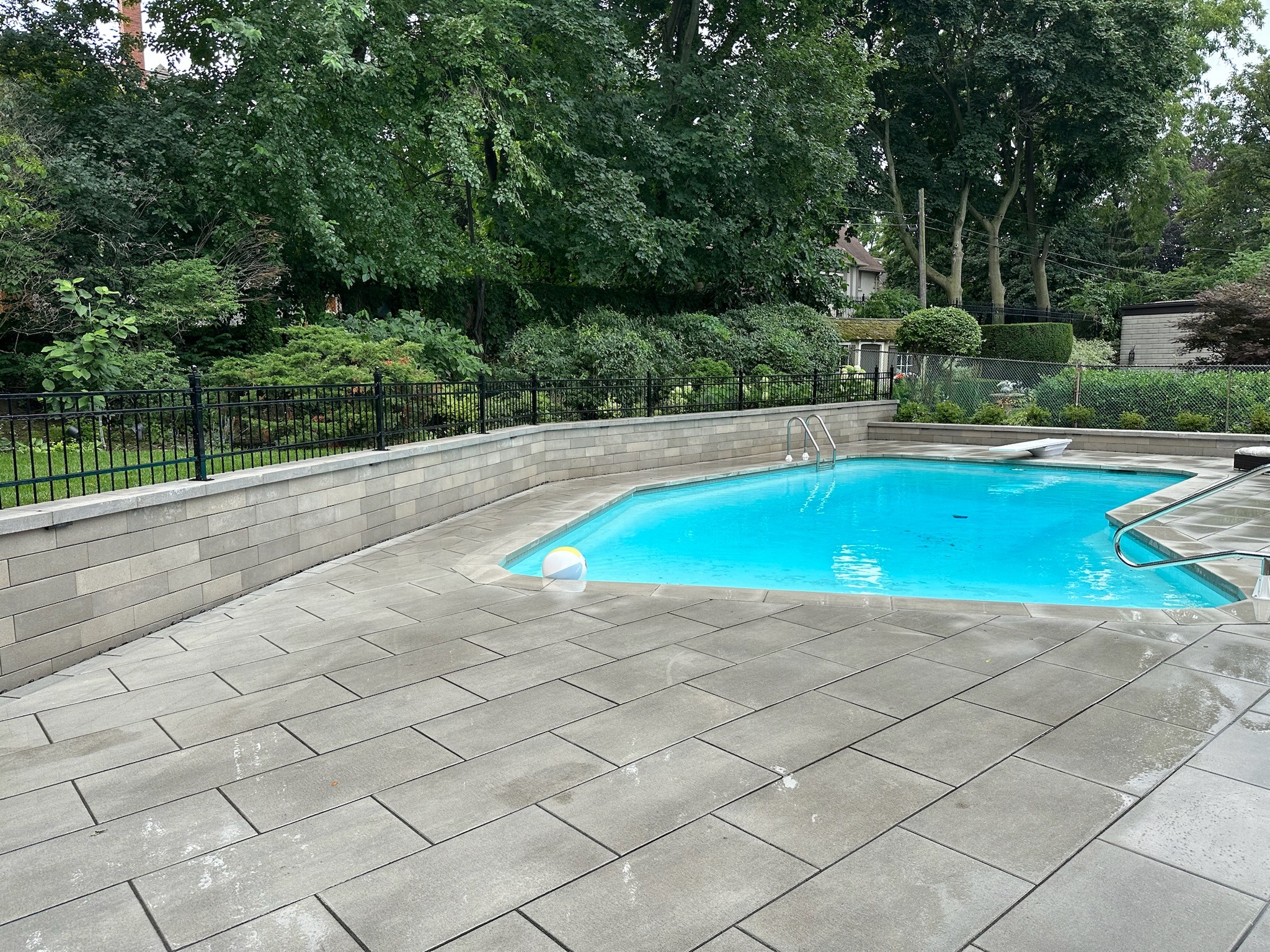 A tranquil backyard swimming pool surrounded by lush greenery and a stone patio, featuring a beach ball and an adjacent residential house.