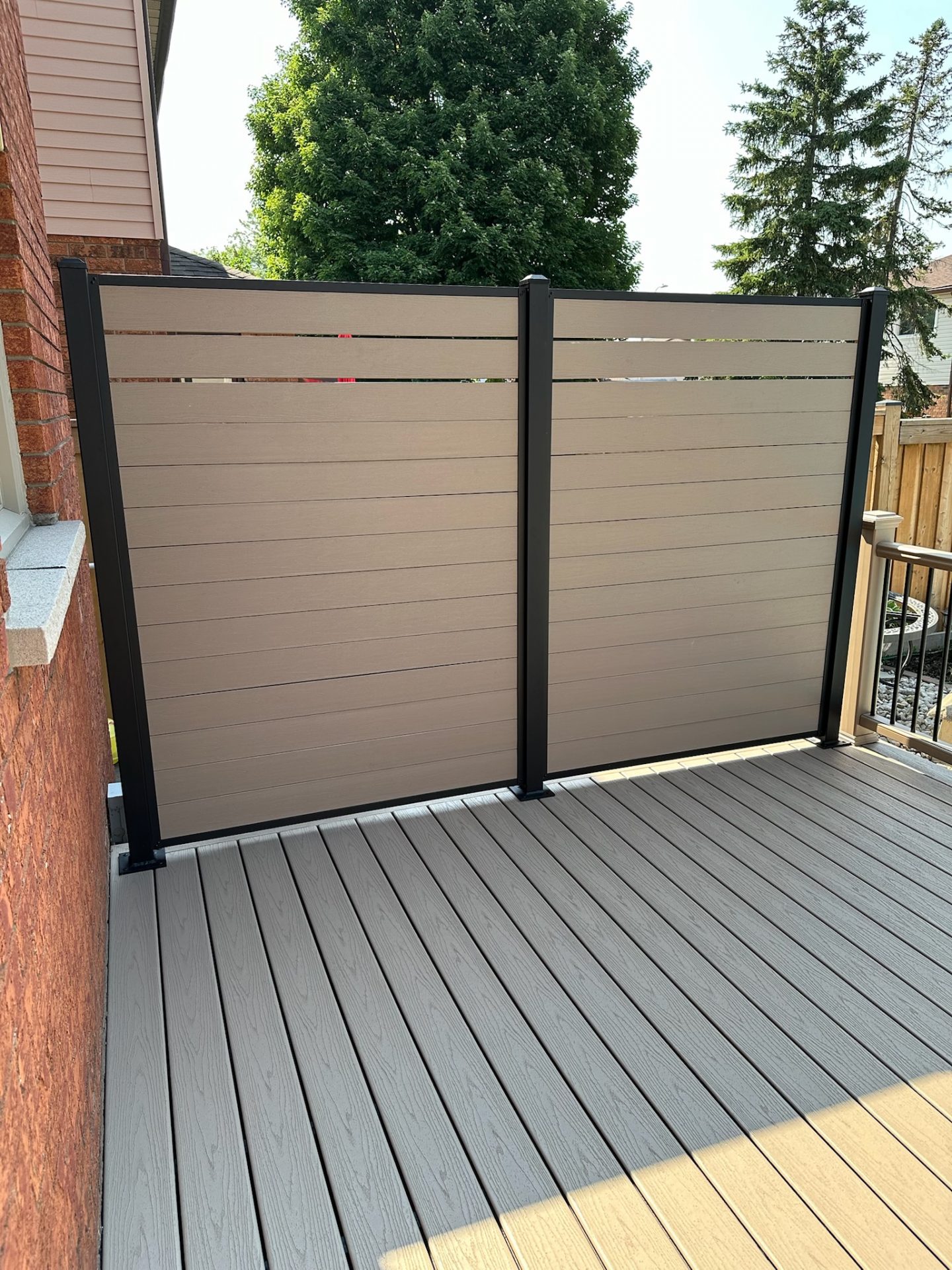 Modern backyard with composite decking, privacy screen, and partial view of red brick house. Lush green trees in the background under a clear sky.