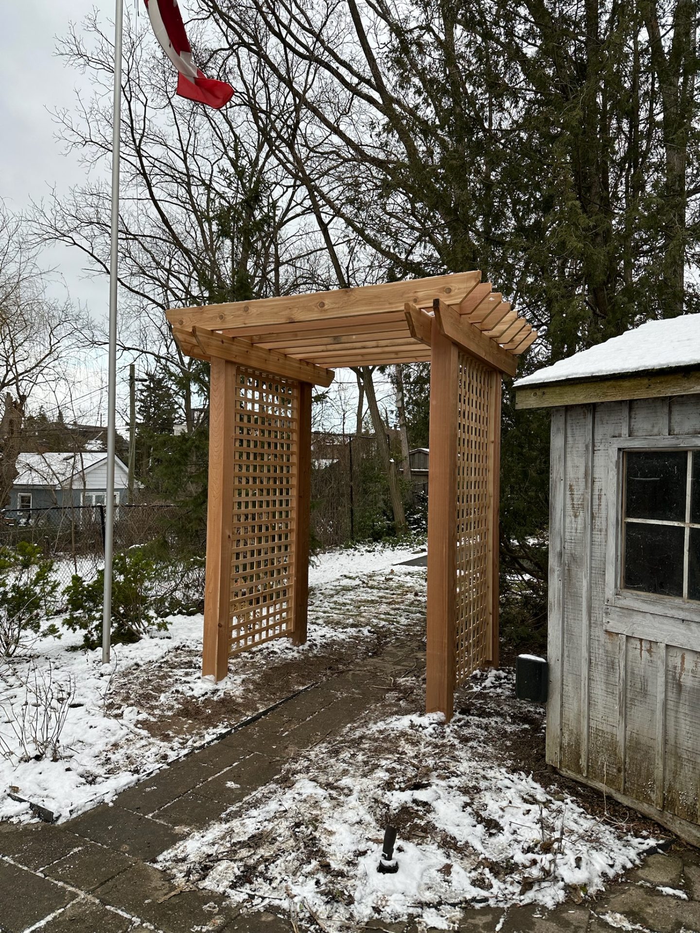 Wooden pergola with lattice panels stands on a snowy path near a weathered shed. Canadian flag flutters on a pole nearby.