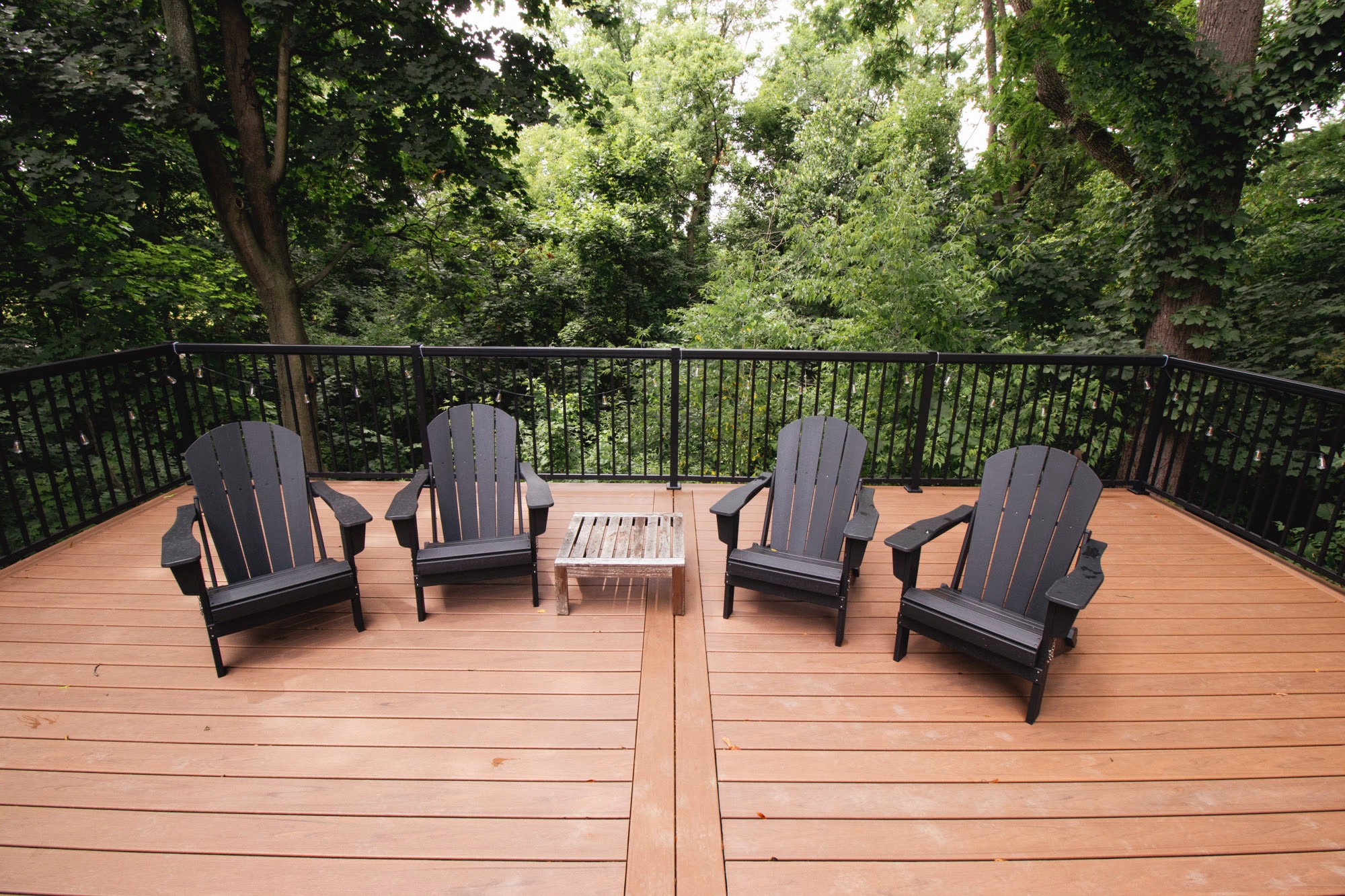 A wooden deck features four Adirondack chairs and a small table, surrounded by lush green trees and a metal railing.