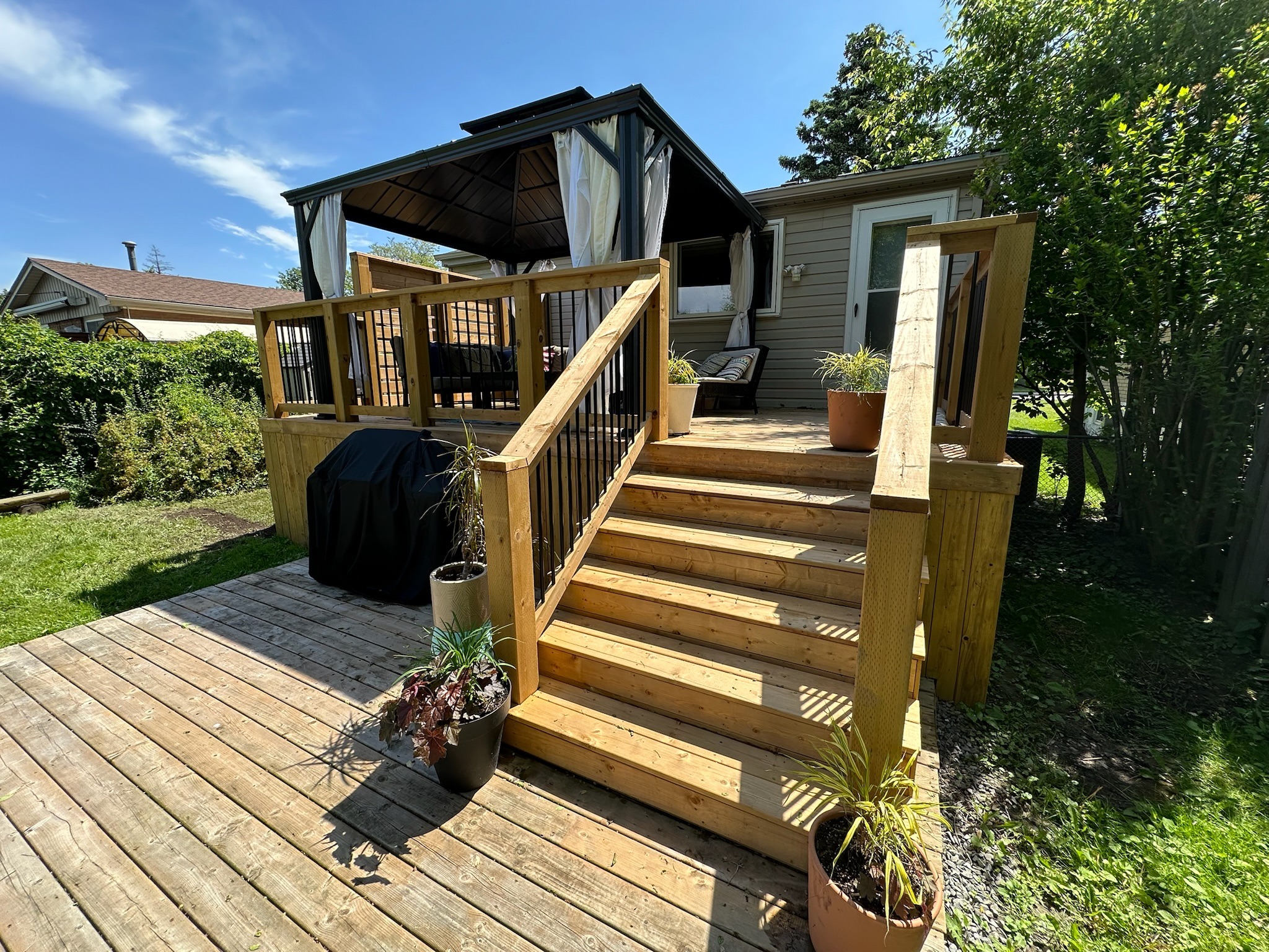 Wooden deck with potted plants and stairs leads to a cozy gazebo-covered porch, surrounded by green shrubbery and lawn under a blue sky.