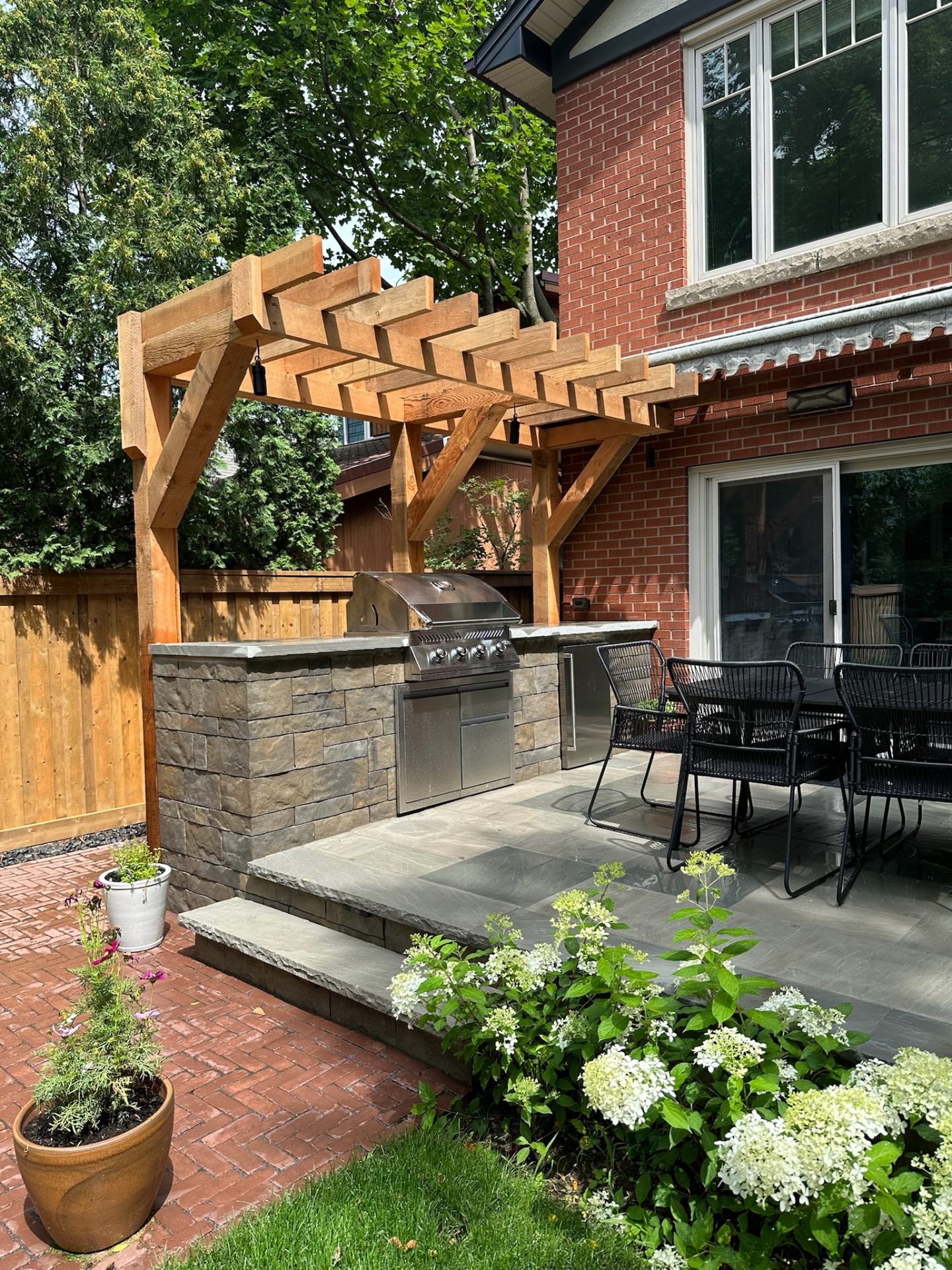 A backyard scene featuring a brick house, stone patio, wooden pergola, grill, dining area, and potted plants, with lush greenery surrounding.