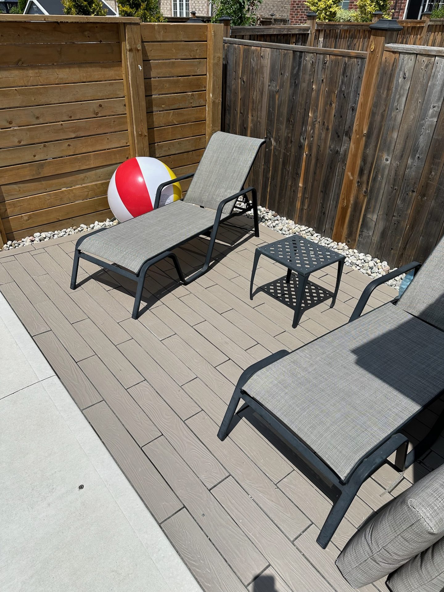Two lounge chairs on a wooden patio with a colorful beach ball, surrounded by a wooden fence under sunny conditions.