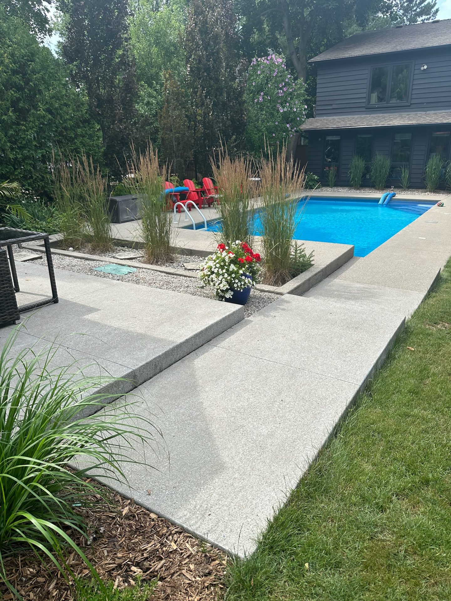 A modern backyard with a swimming pool, surrounding greenery, and red chairs. A two-story house is in the background.