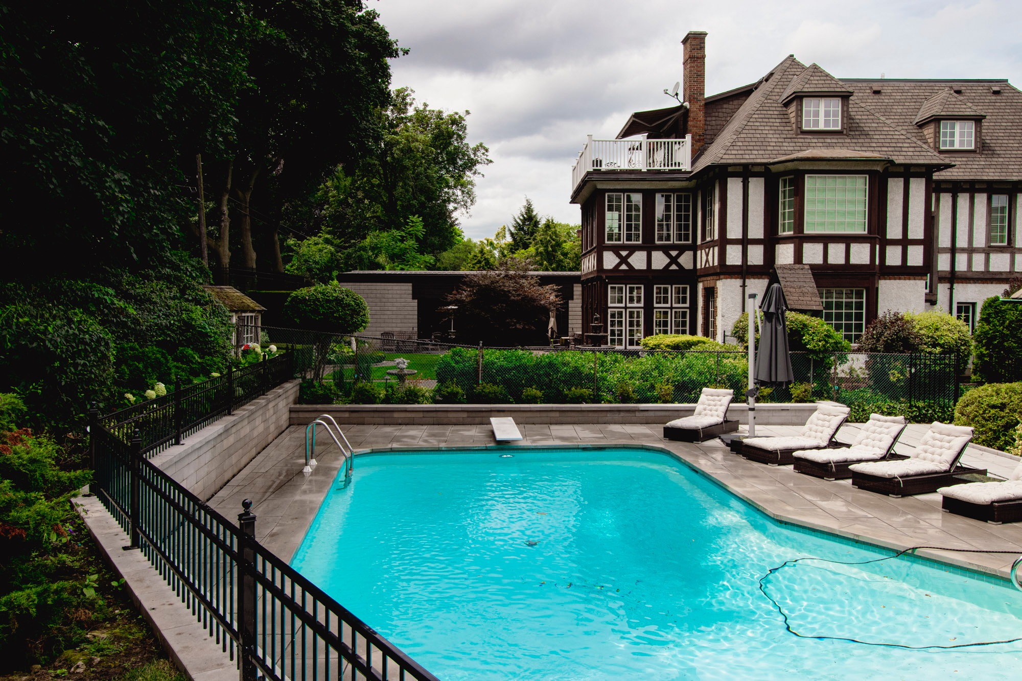 Tudor-style house with pool, loungers, and gardens surrounded by trees. Overcast sky creates a serene atmosphere. No people present.