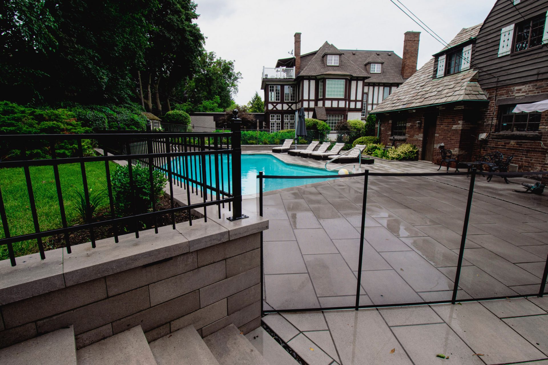 A stone patio surrounds a fenced pool with loungers. A brick house with Tudor-style architecture is in the background, amidst lush greenery.