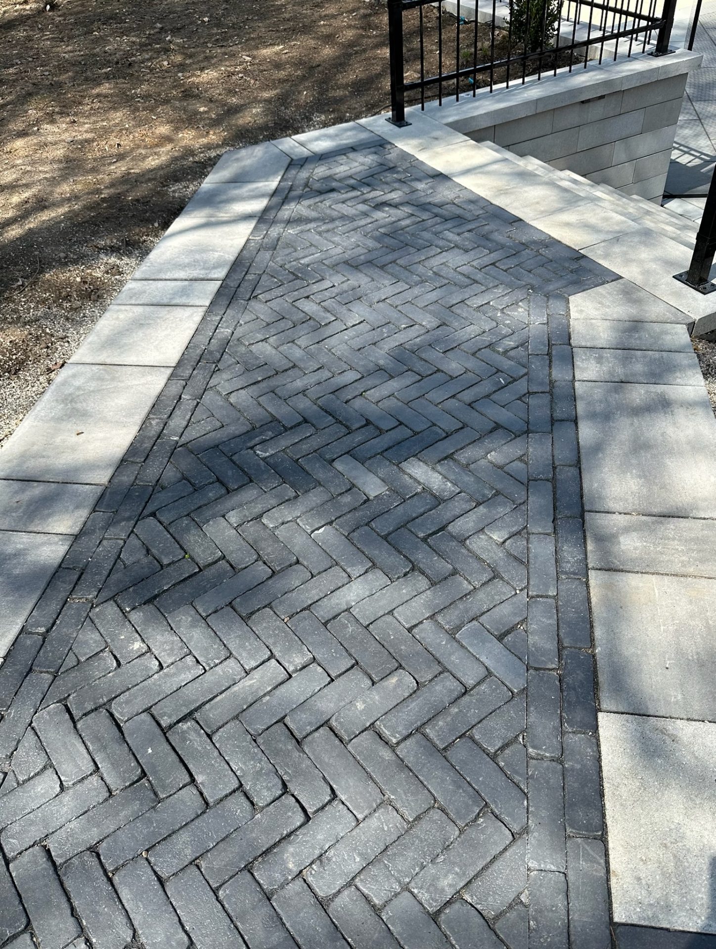 A herringbone-patterned brick pathway with a concrete border, leading to a railing and stairs, surrounded by dirt and shadows.