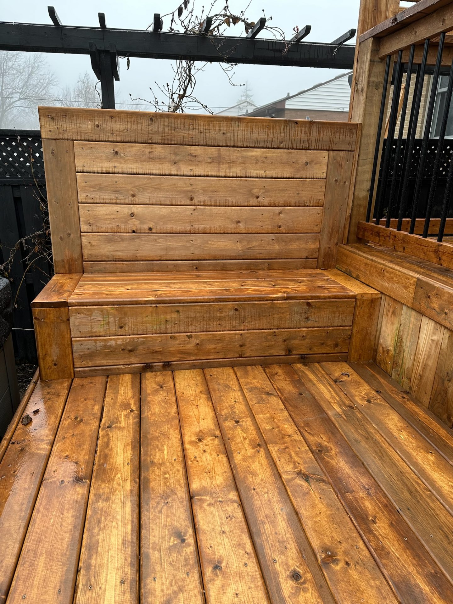 Wooden deck with built-in bench and pergola overhead. Bare branches in the background, creating a rustic and serene outdoor seating area.