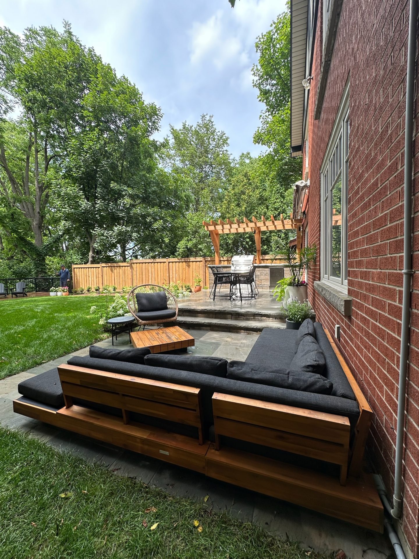 A cozy backyard features a wooden patio set, brick exterior, lush trees, and greenery. A person stands beside a wooden fence.