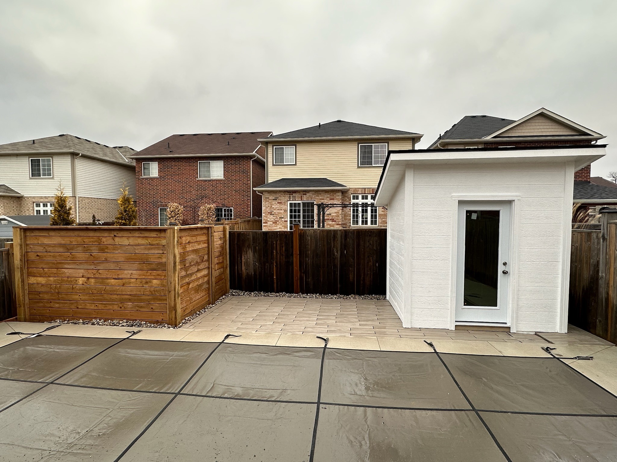 Suburban backyard view with a covered pool, wooden fence, and small white shed. Overcast sky and neighboring houses in the background.