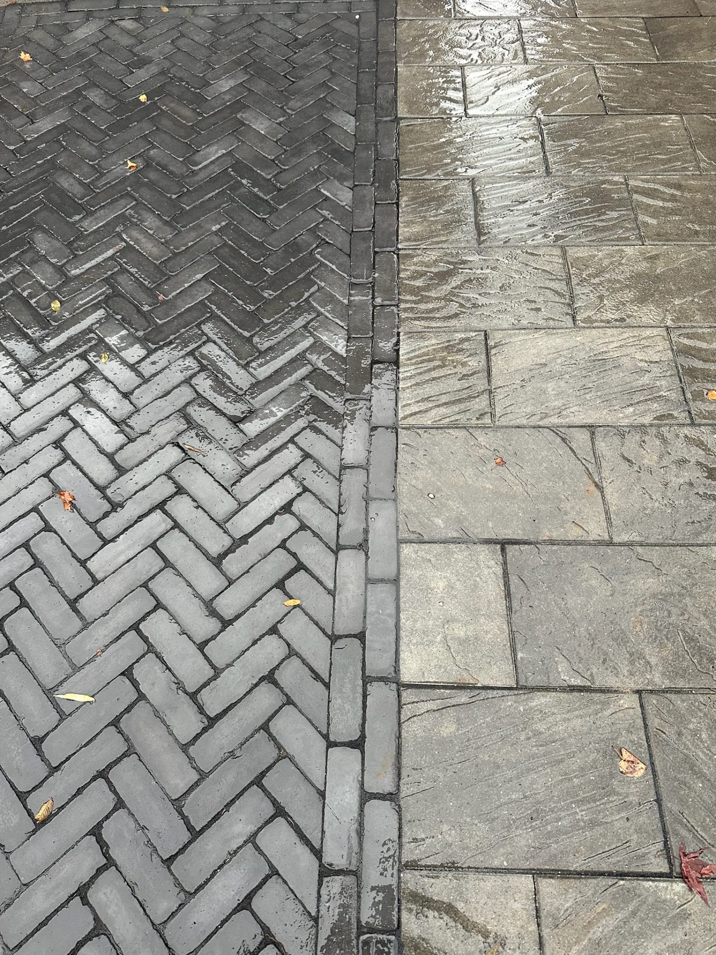 Wet interlocking herringbone and square cobblestones, with scattered leaves, create a textured contrast. No people or landmarks are visible in this sidewalk scene.