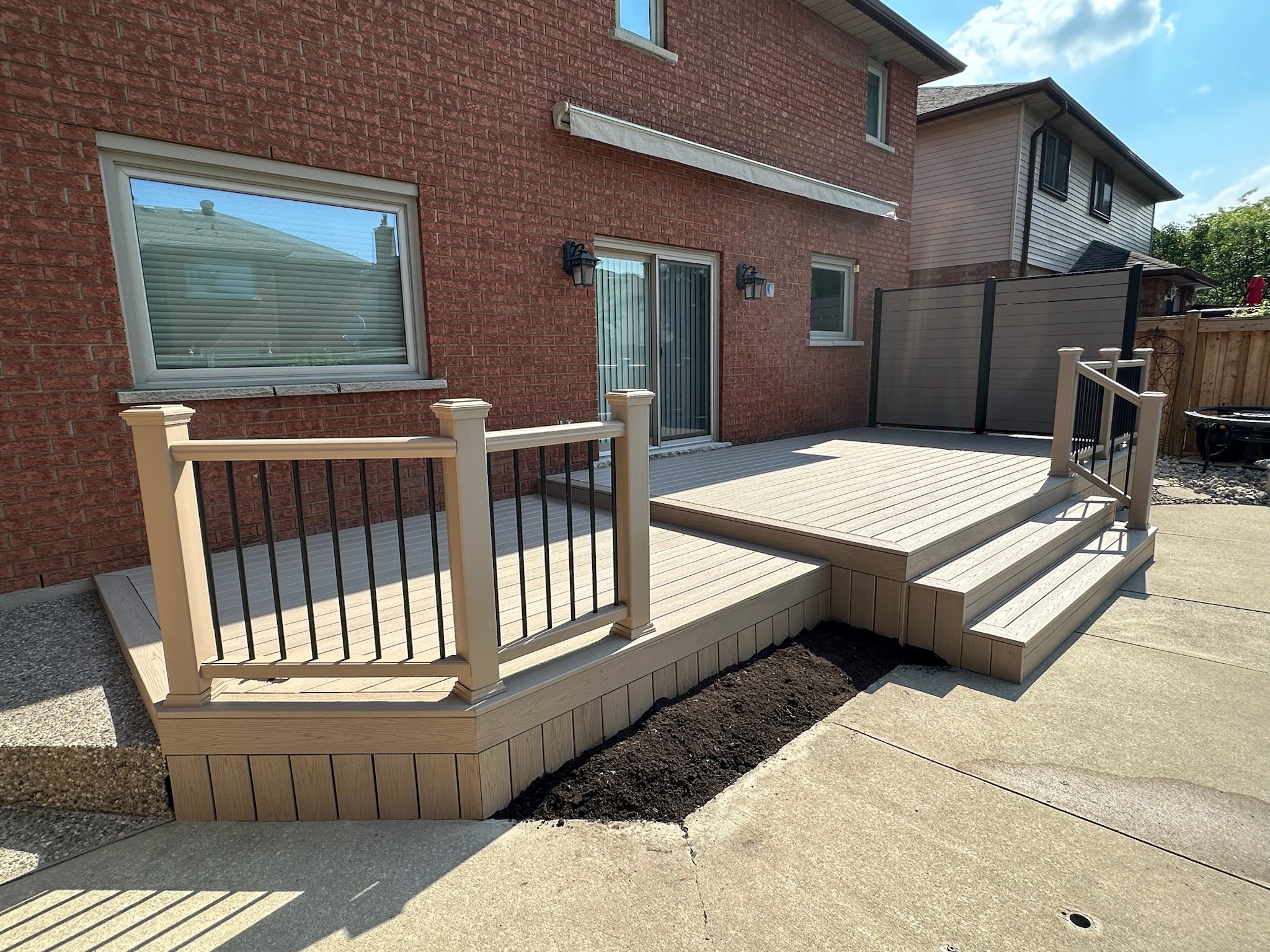A modern backyard deck features multi-level wooden steps and railings, adjacent to a brick house under a bright, partly cloudy sky.