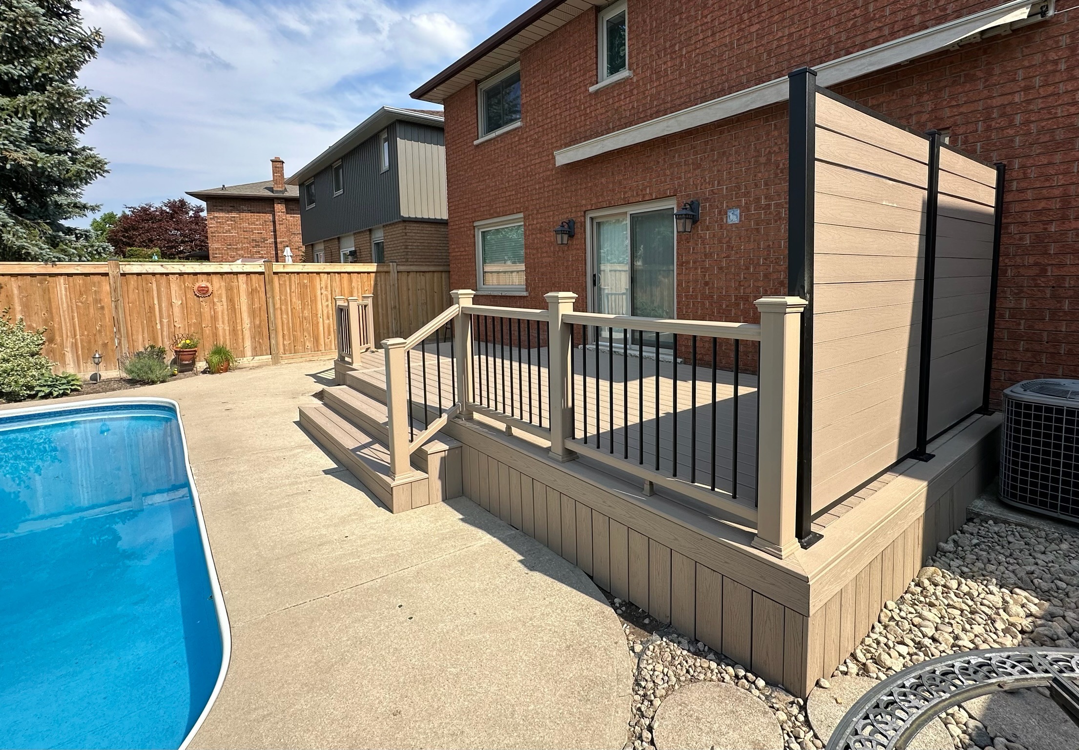 A backyard with a deck, pool, and wooden privacy fence. Red brick house, concrete pathway, and landscaped with plants and stones.