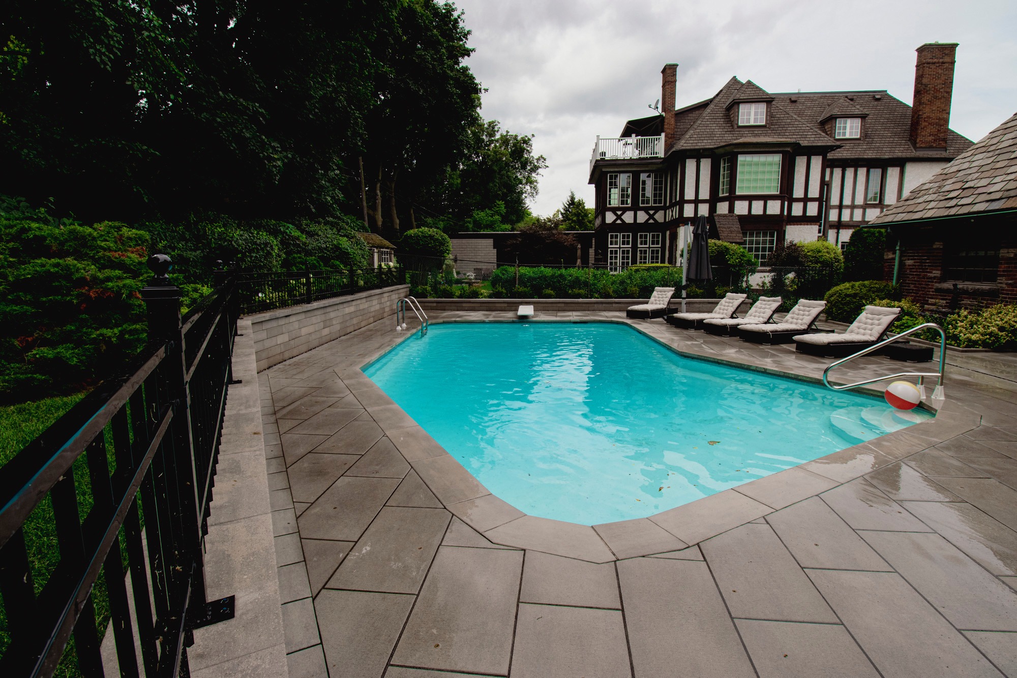 A private backyard view features a clear blue pool, lounge chairs, lush greenery, and a large traditional house under a cloudy sky.