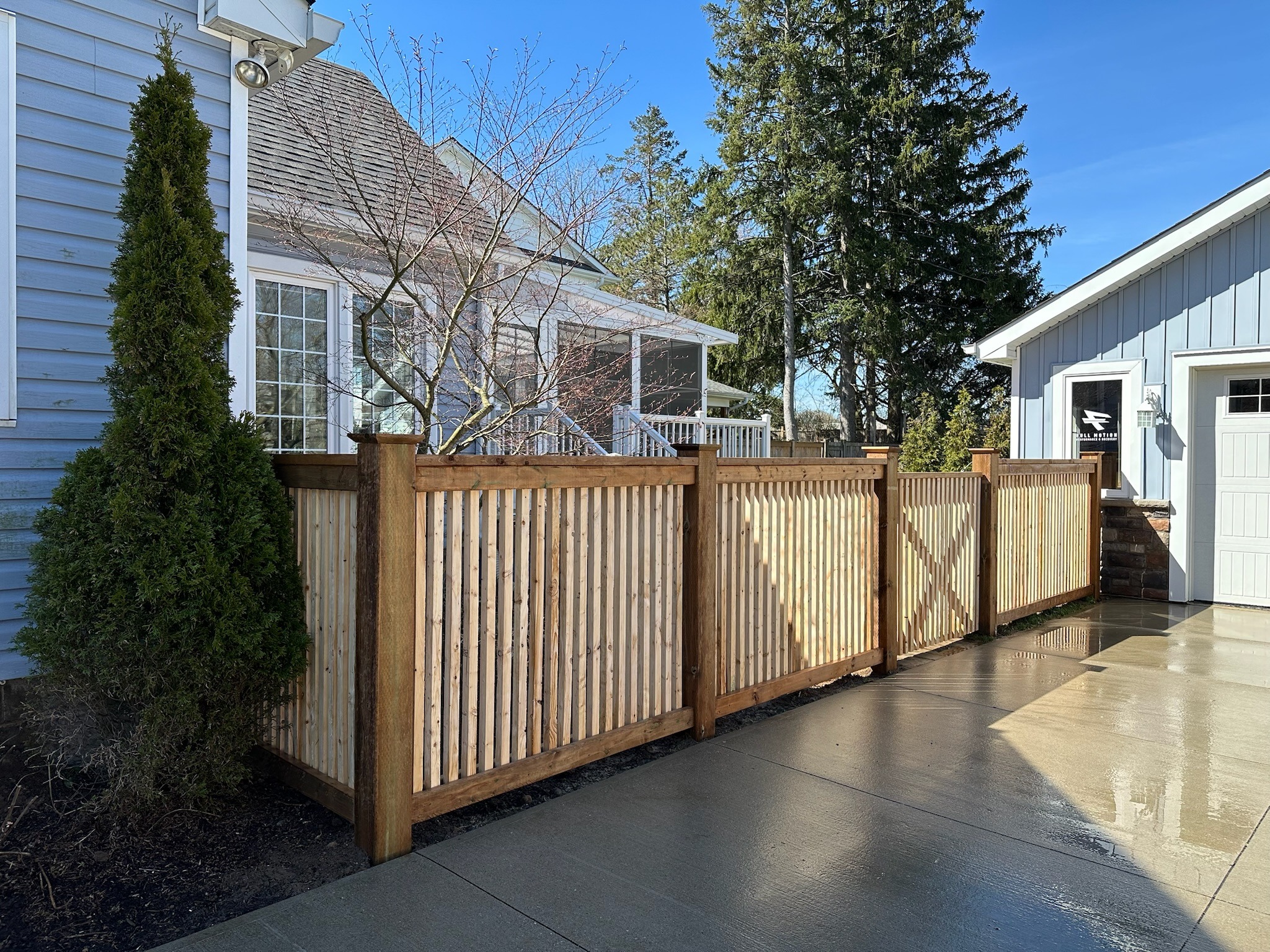A wooden fence separates two blue houses with large windows, surrounded by trees. The ground is wet, reflecting sunlight.