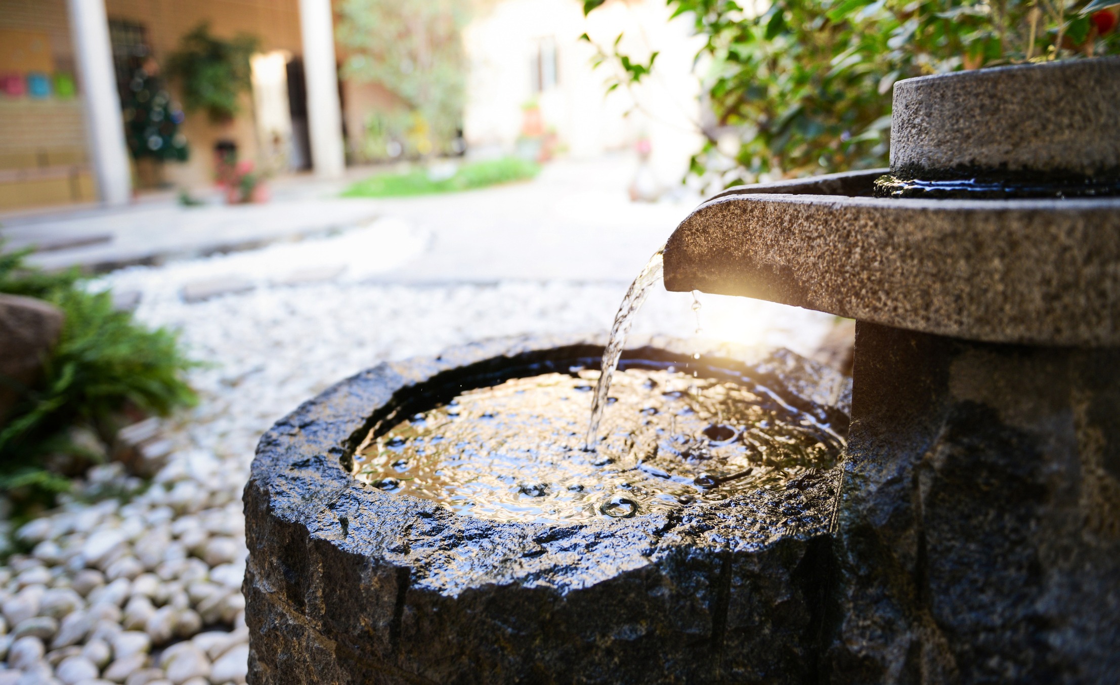 A stone fountain gently pours water in a serene garden courtyard, surrounded by pebbles and lush greenery, bathed in warm sunlight.