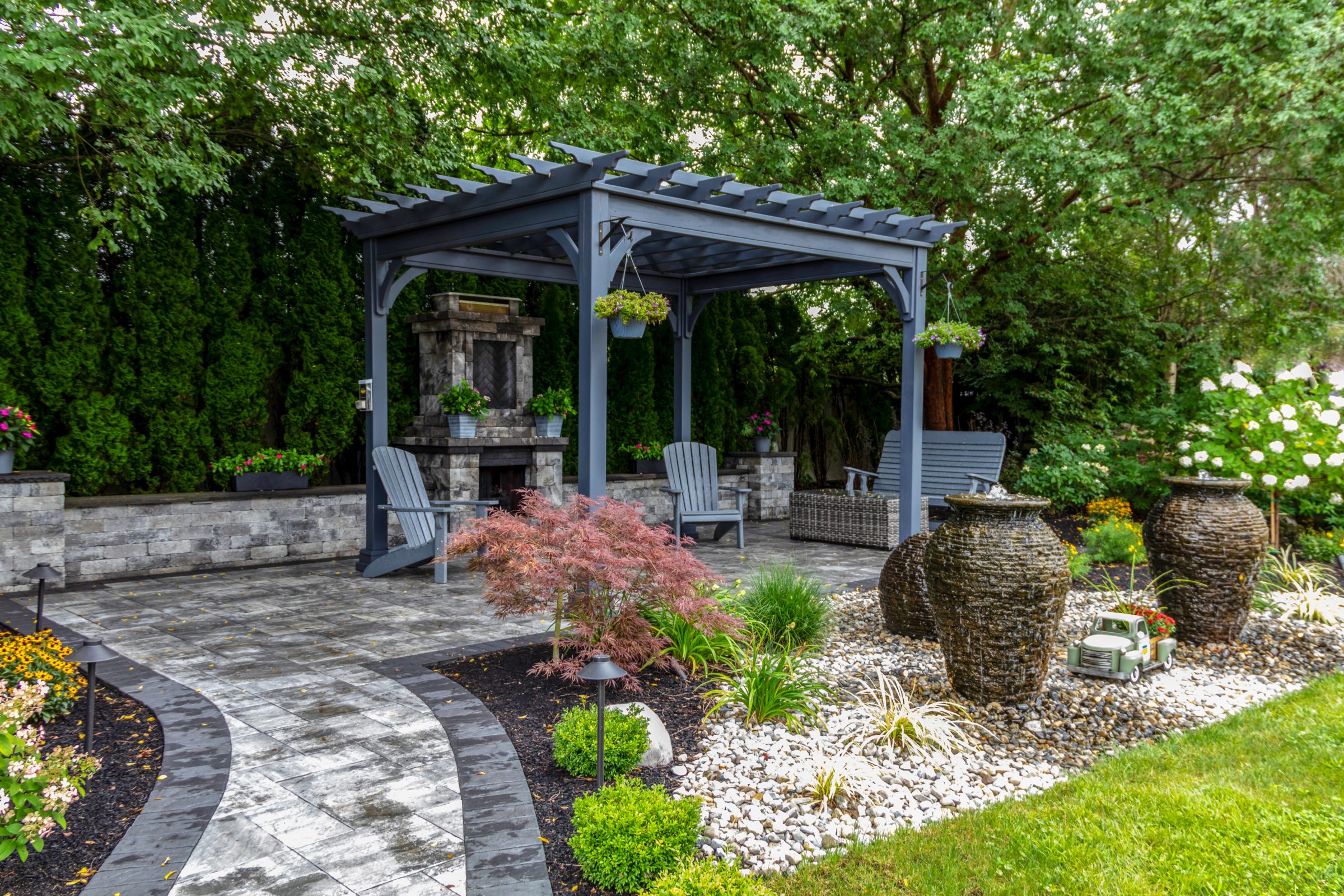A serene garden with pergola, chairs, fireplace, and decorative pots. Lush greenery and pebble landscaping create a tranquil outdoor retreat.