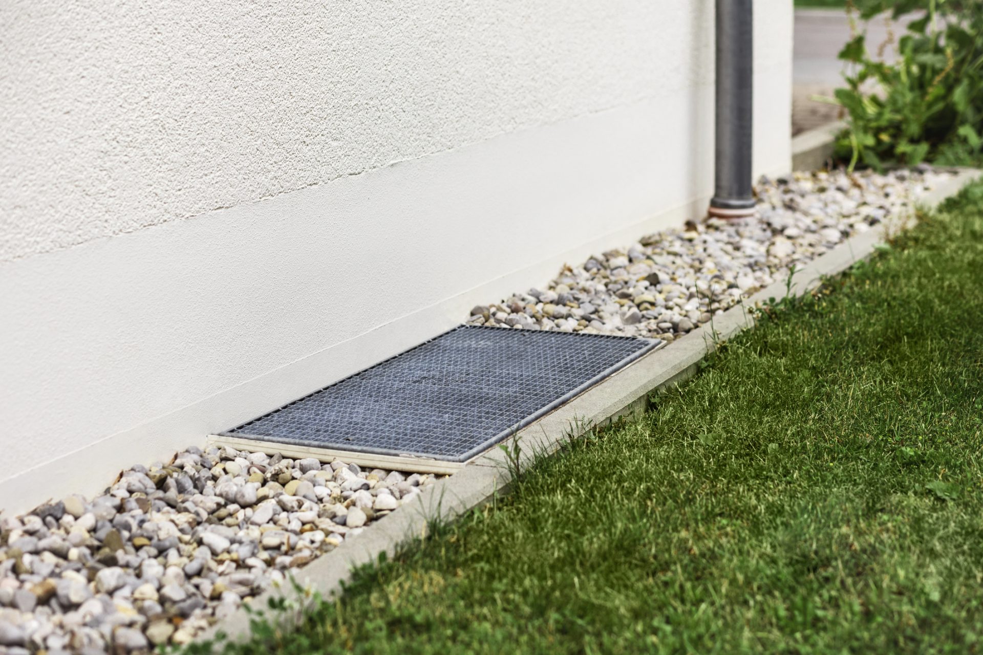 A white building wall with a small metal grate, a rocky border, and adjacent green grass. No recognizable landmarks or buildings are visible.
