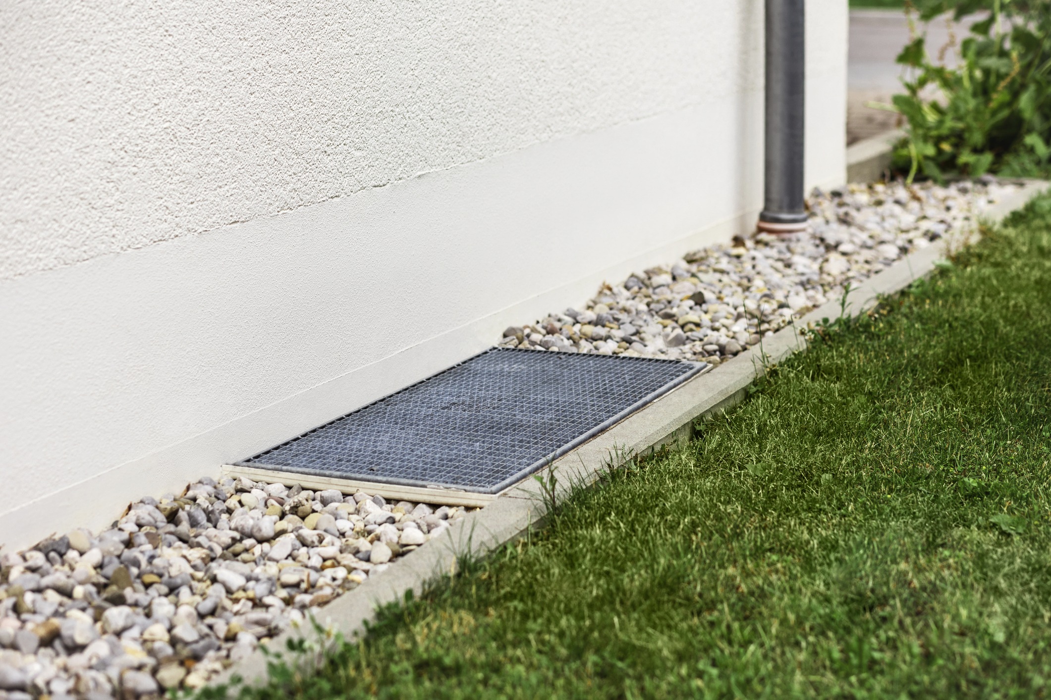 A white building wall with a small metal grate, a rocky border, and adjacent green grass. No recognizable landmarks or buildings are visible.