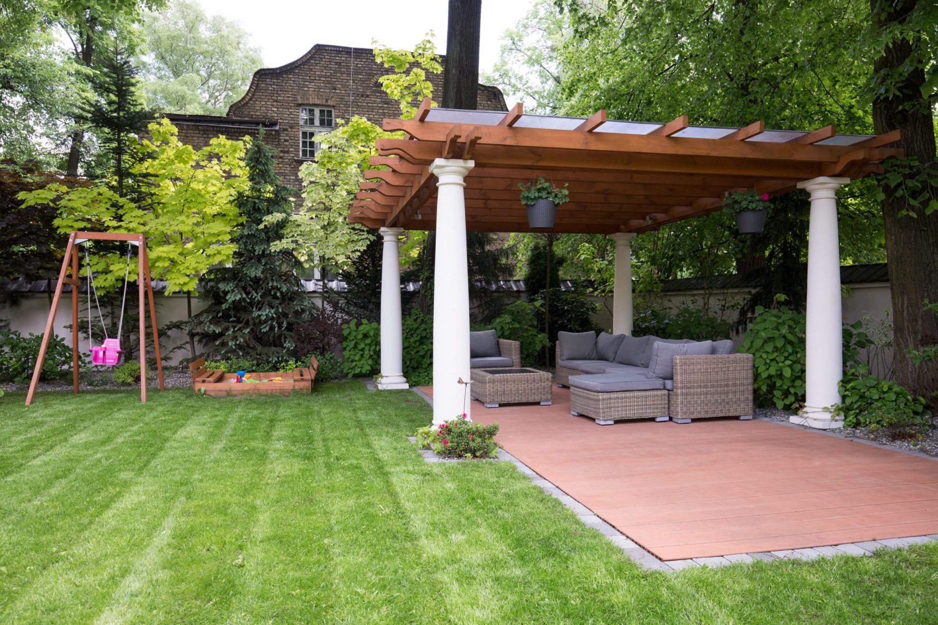A garden features a wooden pergola with seating, a swing, sandbox, and vibrant greenery, adjacent to a historic brick building.
