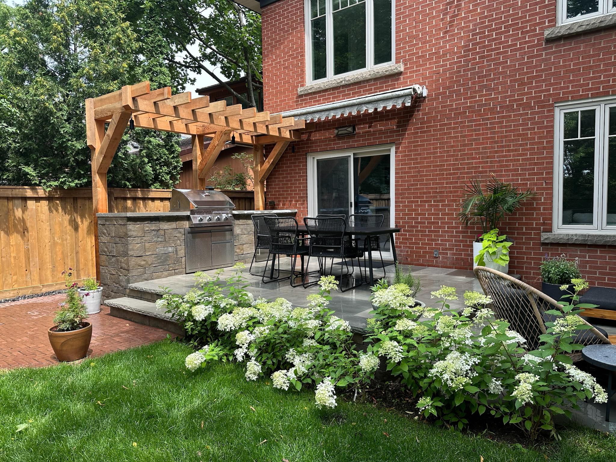 A backyard outdoor kitchen and dining area with a stone-built barbecue island, wooden pergola, patio furniture, and blooming hydrangeas beside a red brick house.