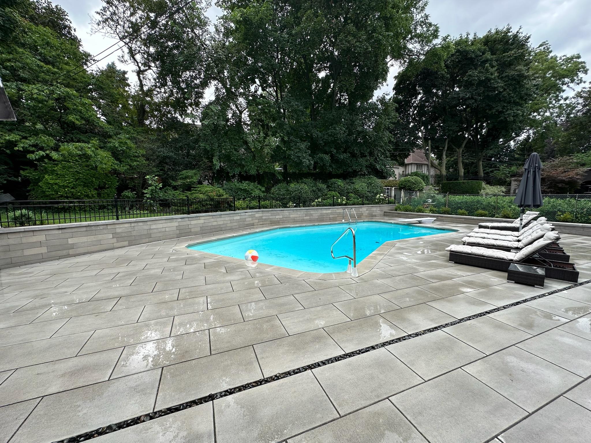 A backyard pool bordered by wet gray pavers after rainfall, with stacked lounge cushions and a backdrop of tall trees and greenery.