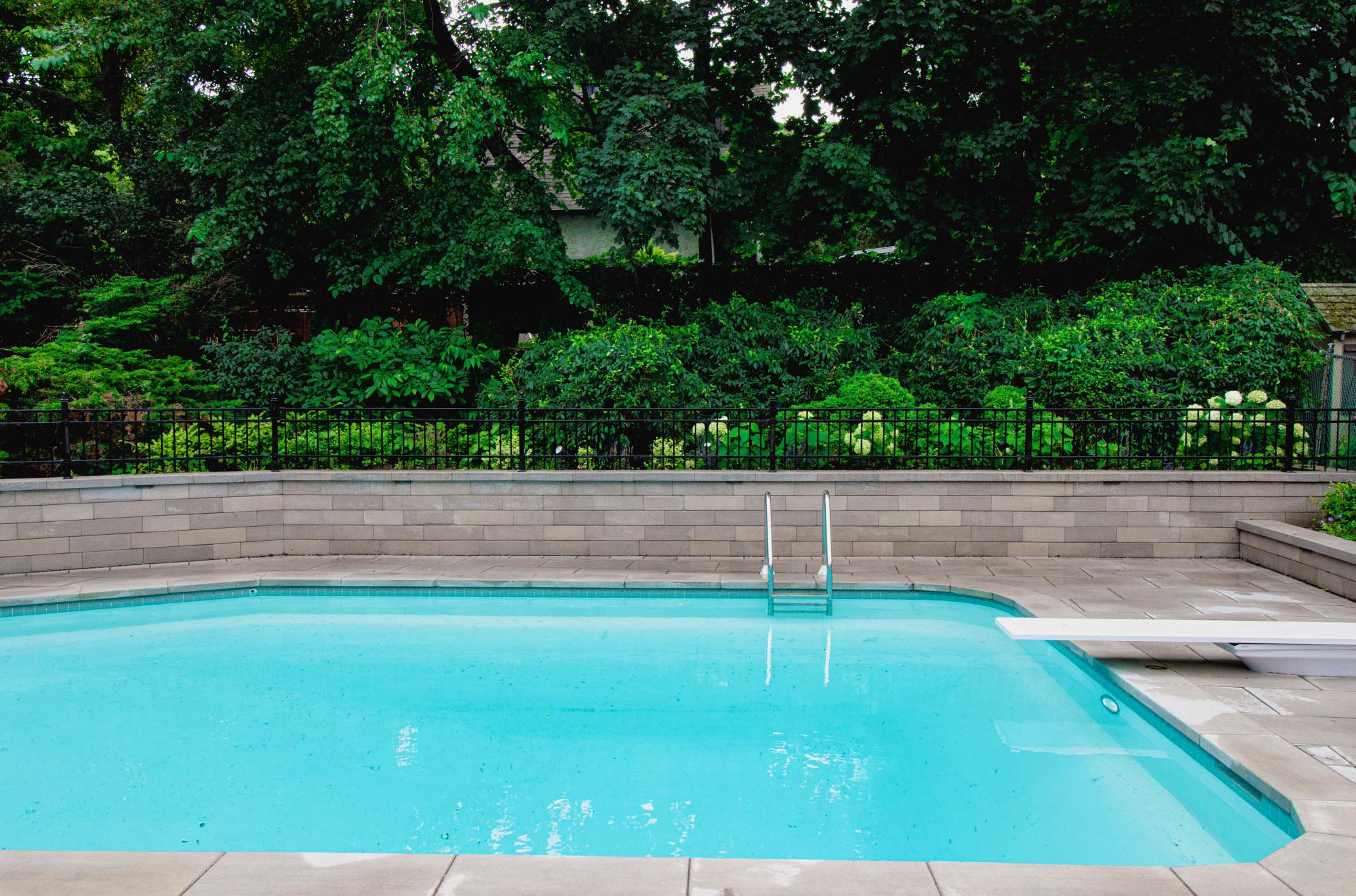 A backyard swimming pool surrounded by stone pavers, with a retaining wall and dense greenery behind a black metal fence.