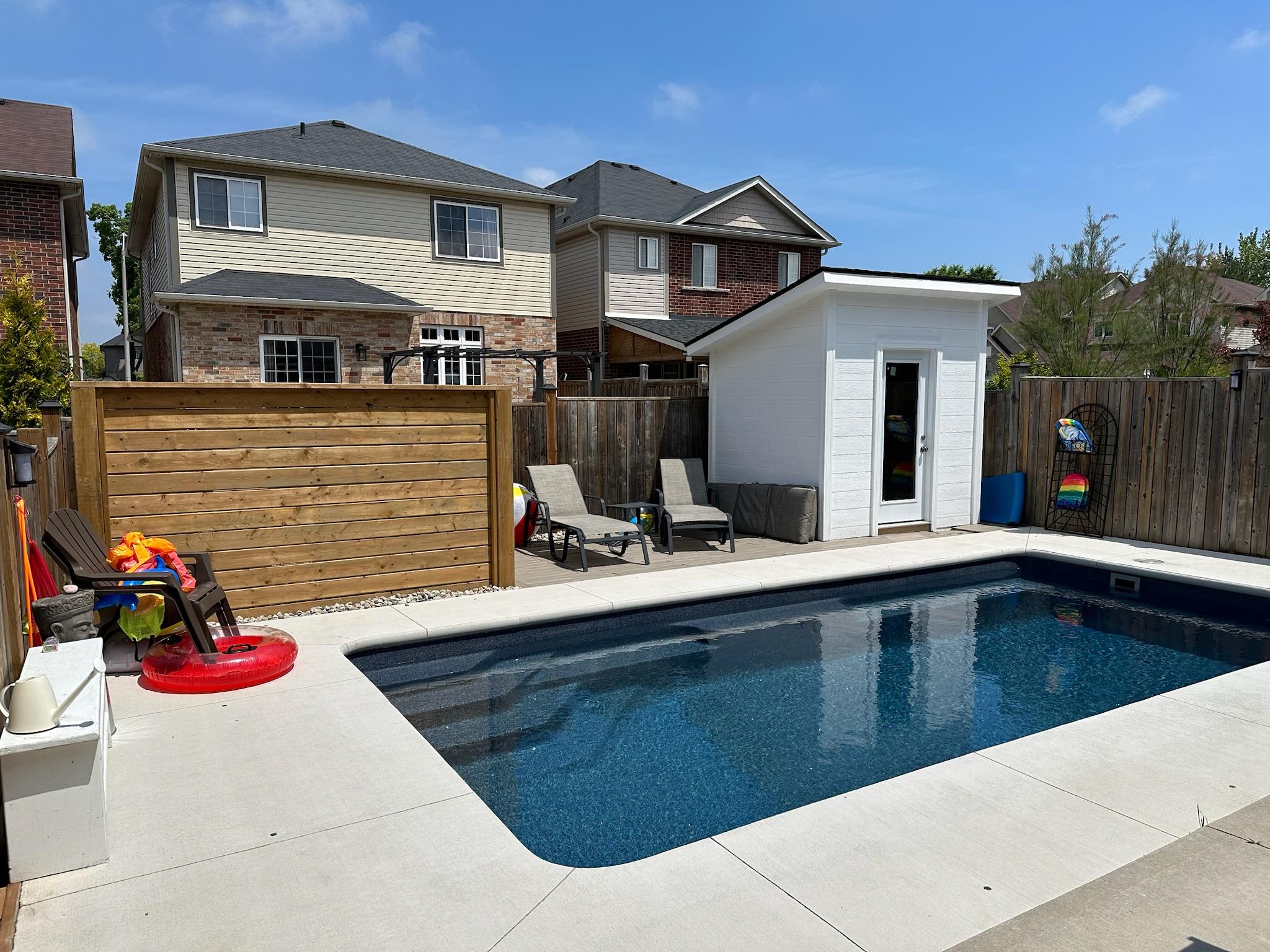A backyard swimming pool with a concrete deck, wooden privacy screen, lounge chairs, and a small white pool house.