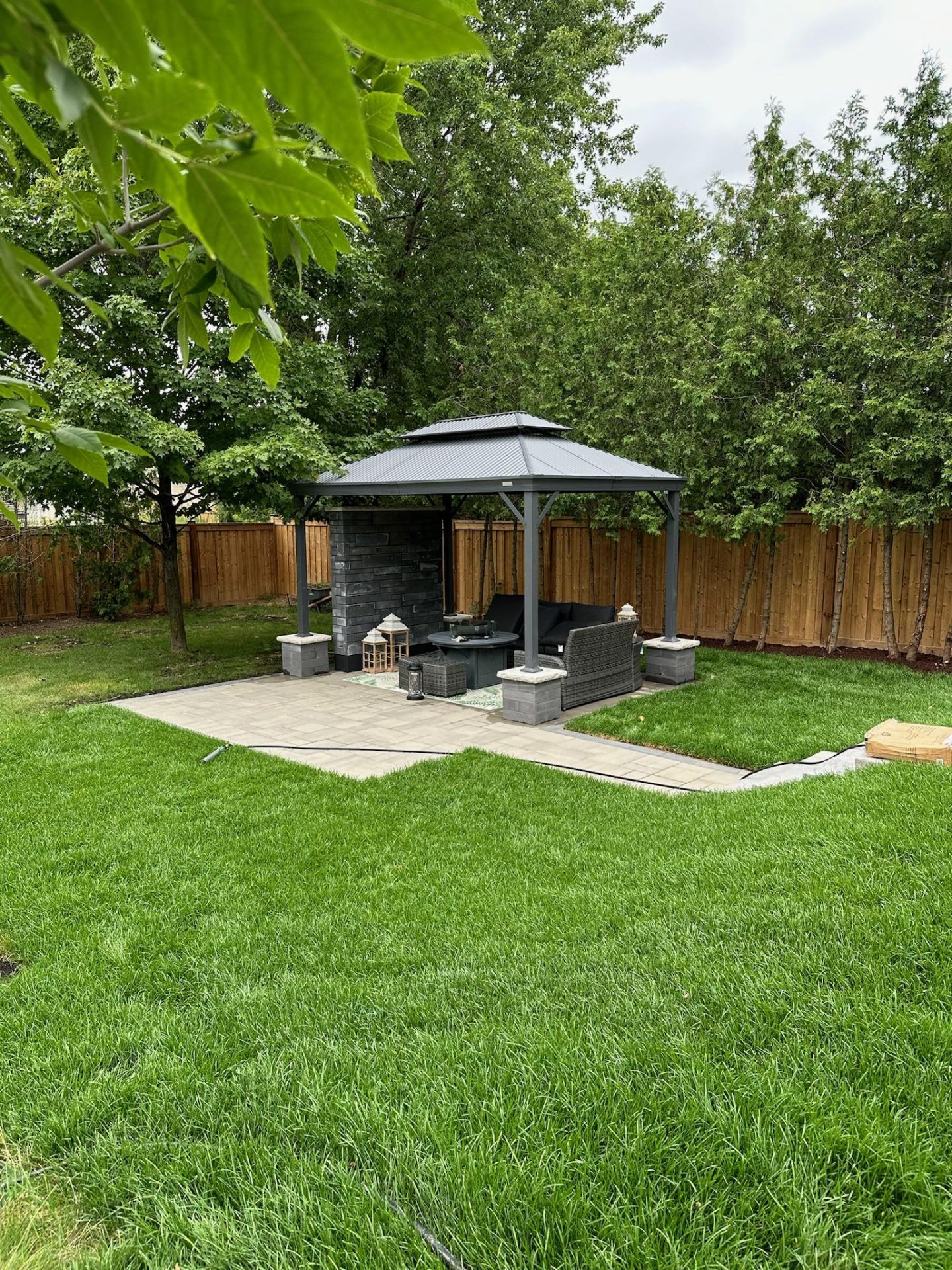 A backyard with a grey gazebo, stone fireplace feature, wicker seating, and lush green lawn surrounded by wooden fencing.