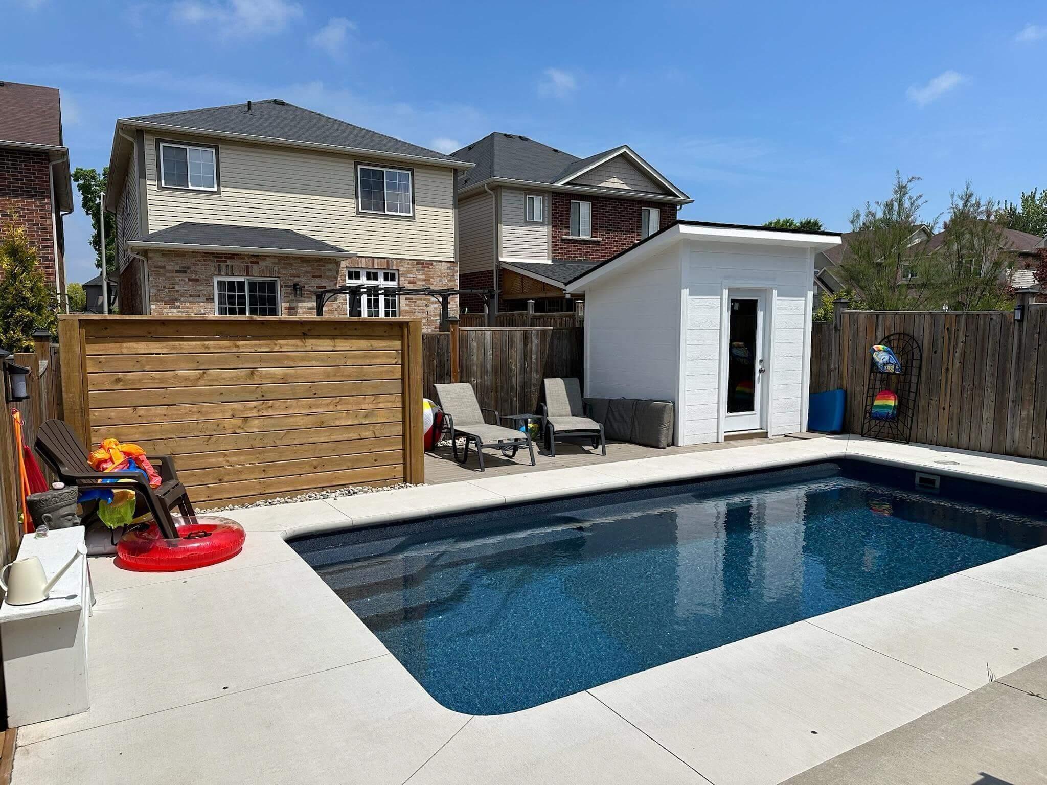 A backyard with a rectangular in-ground pool, concrete pool deck, lounge chairs, wooden privacy panel, and a small white pool house.