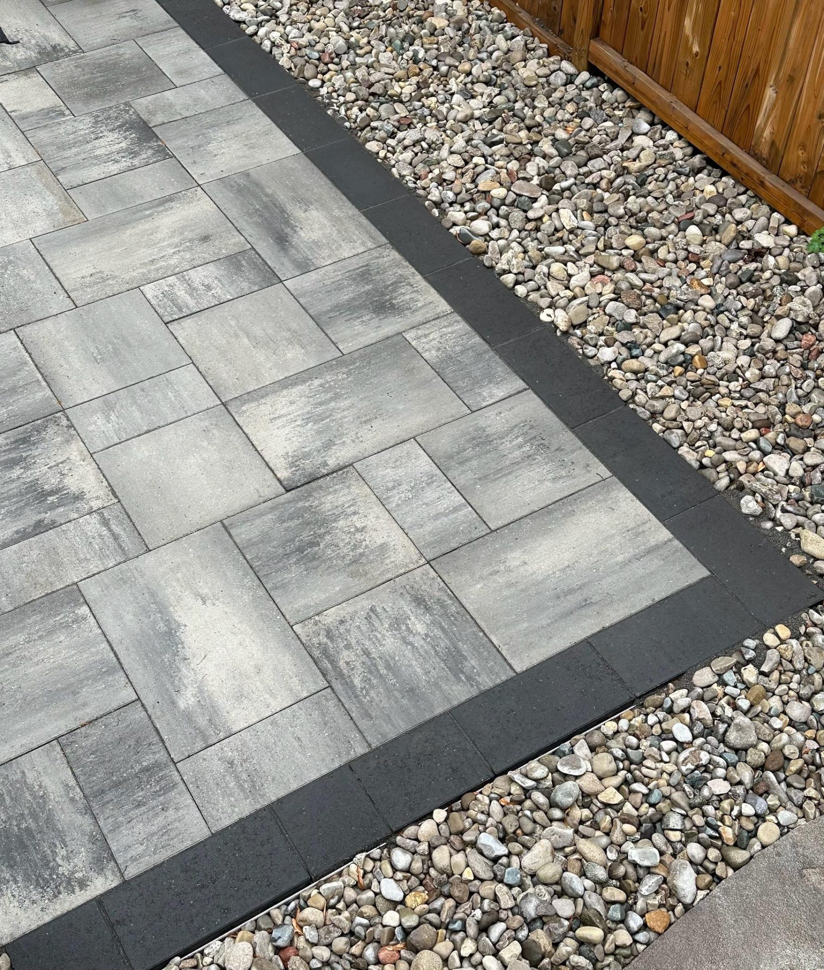 A close-up of grey interlocking pavers bordered with dark stone edging beside a river rock drainage strip along a wooden fence.