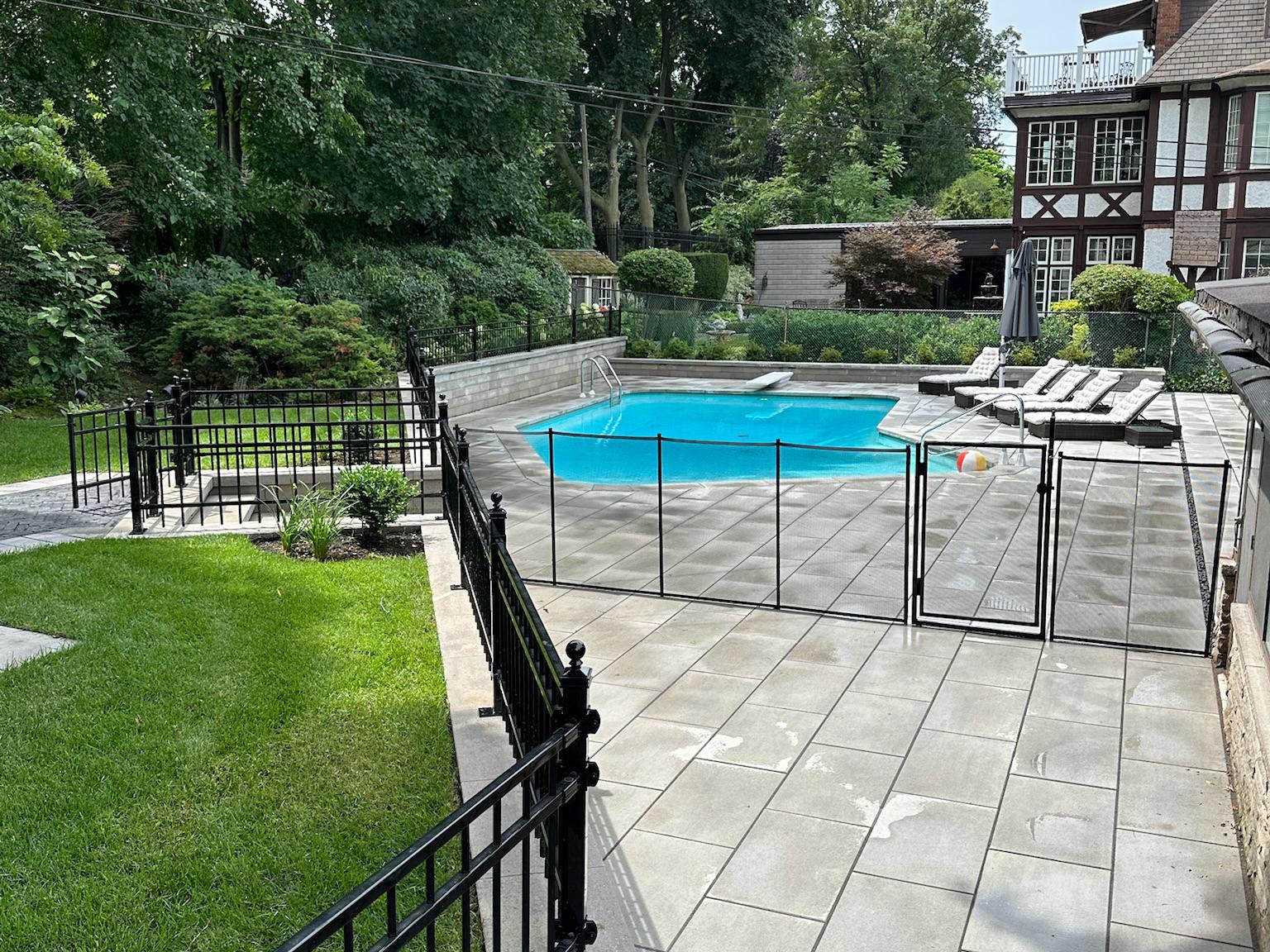 A fenced-in backyard swimming pool surrounded by light gray pavers, with lounge chairs at one end and mature trees and shrubs around the perimeter.