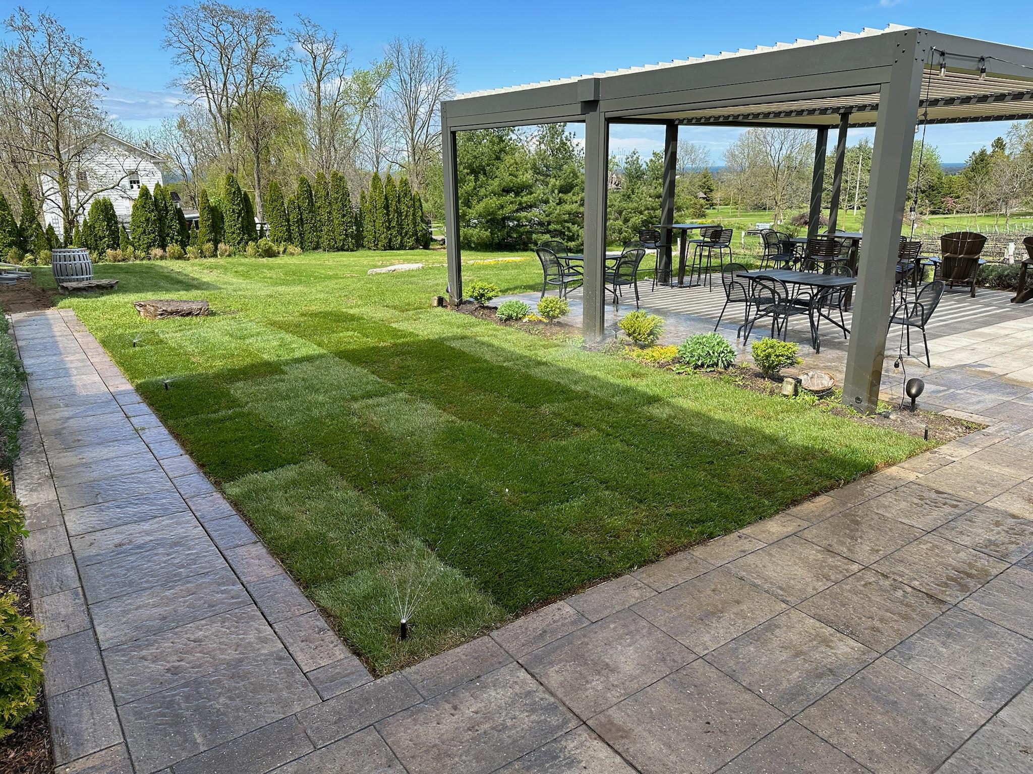 A large backyard with a metal pergola over a dining area, bordered by fresh green sod, planting beds, and stone walkways.
