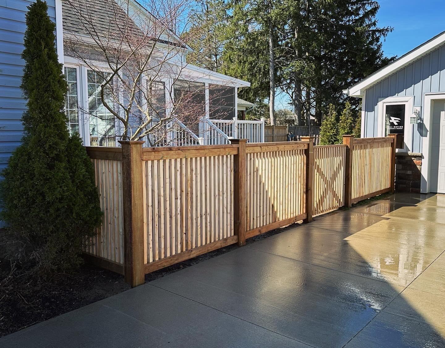 A modern wooden fence with vertical slats and sturdy posts bordering a paved driveway beside residential homes.