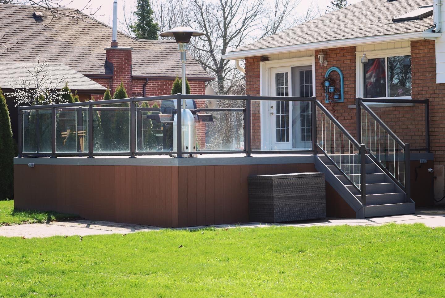 A raised composite deck with glass railing, steps leading to a patio, a heater, and a barbecue beside a brick house.