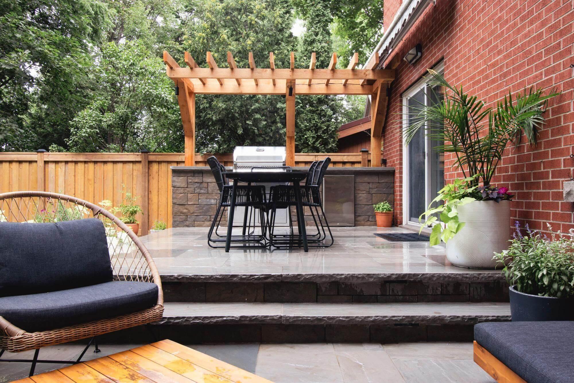 A raised stone patio with a wooden pergola, outdoor dining set, built-in grill island, and potted plants beside a brick house.