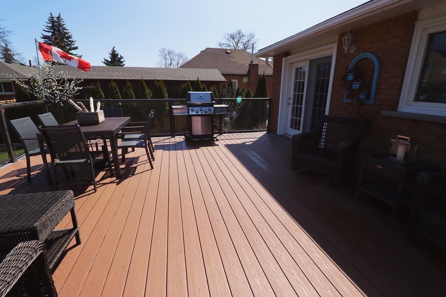 A spacious composite deck with black-framed glass railing, outdoor dining set, wicker seating, and a barbecue next to a brick home.