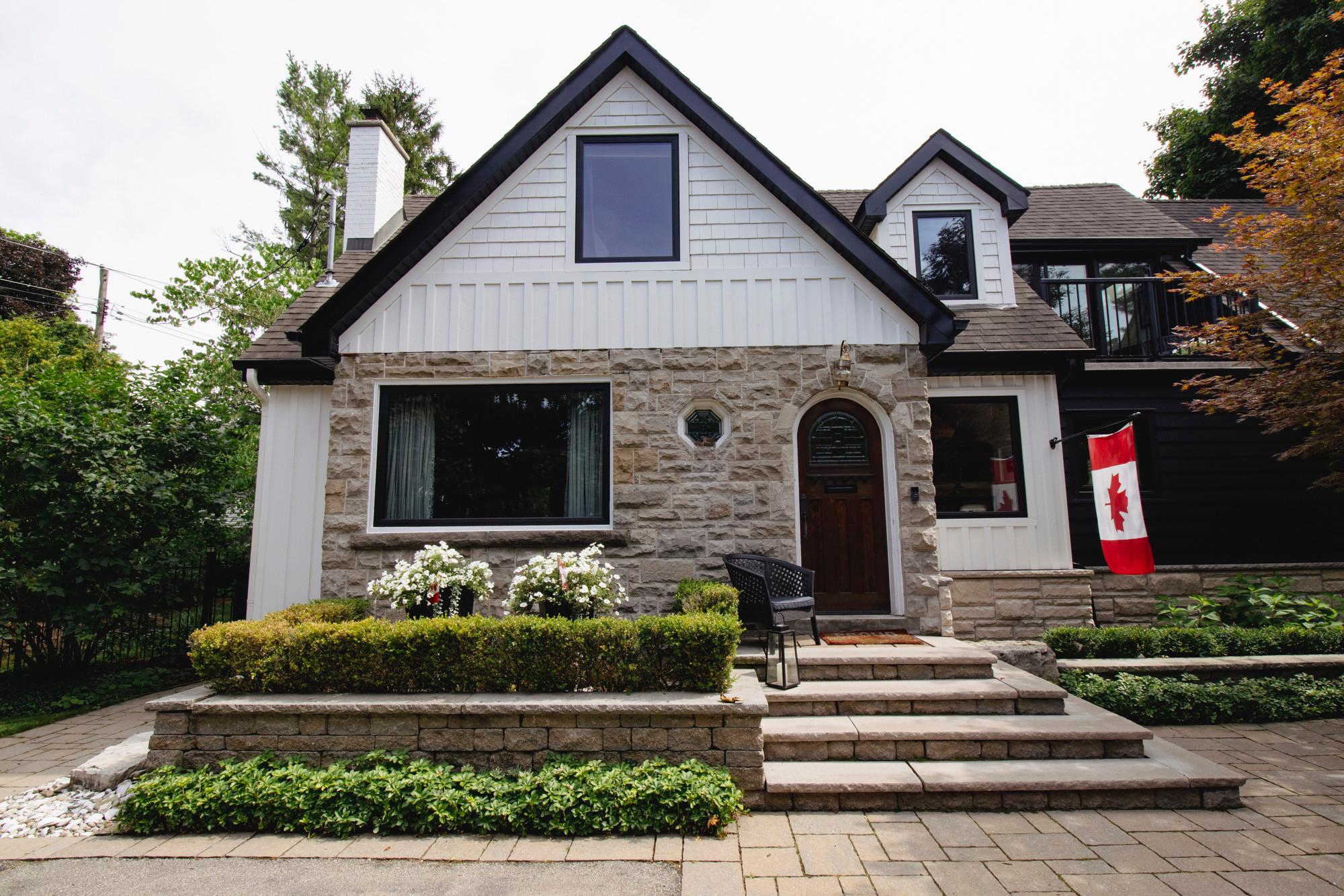 A stone-clad home with white siding accents, tiered garden beds, trimmed hedges, white flowers, and a Canadian flag by the front entrance.