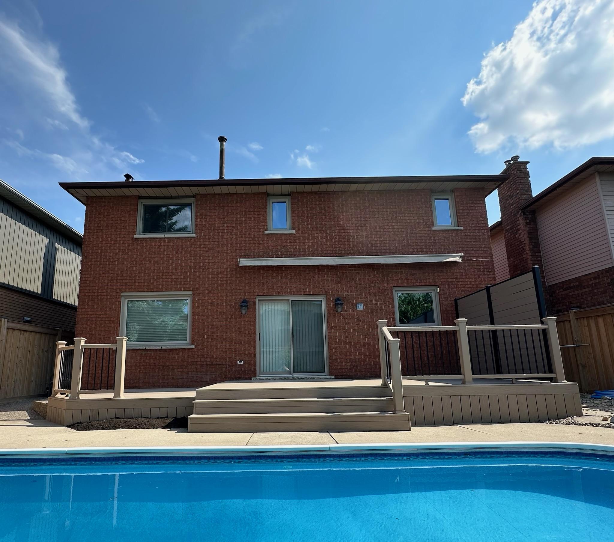 A wide stamped concrete pool deck with a large composite deck and privacy screen attached to a two-story brick home.