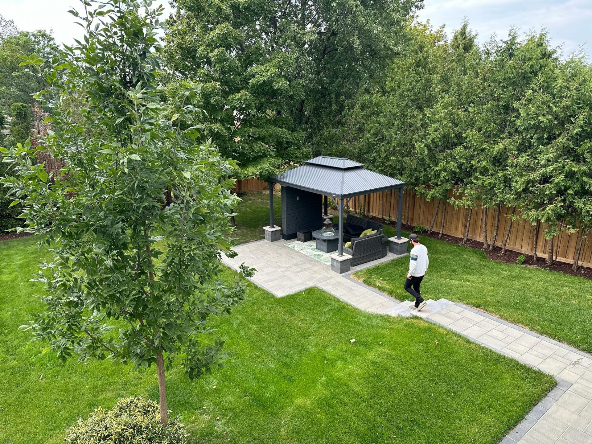 An aerial view of a landscaped backyard featuring a paved walkway leading to a covered gazebo with outdoor seating, surrounded by green lawn and a wooden fence.