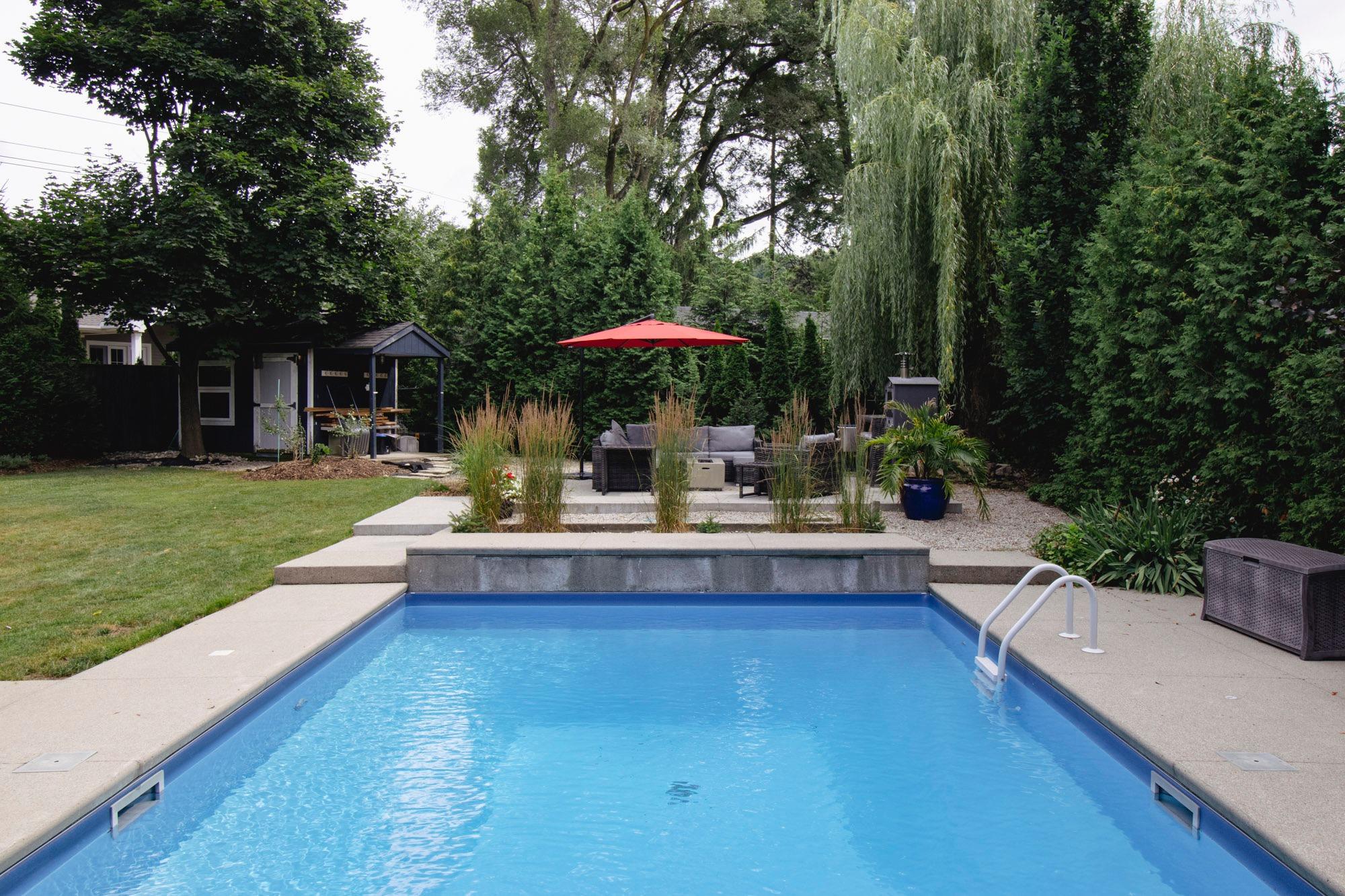 A backyard swimming pool with light blue water bordered by a concrete deck, leading to a raised patio area with tall ornamental grasses, outdoor seating, and a red umbrella, all surrounded by dense greenery and mature trees.