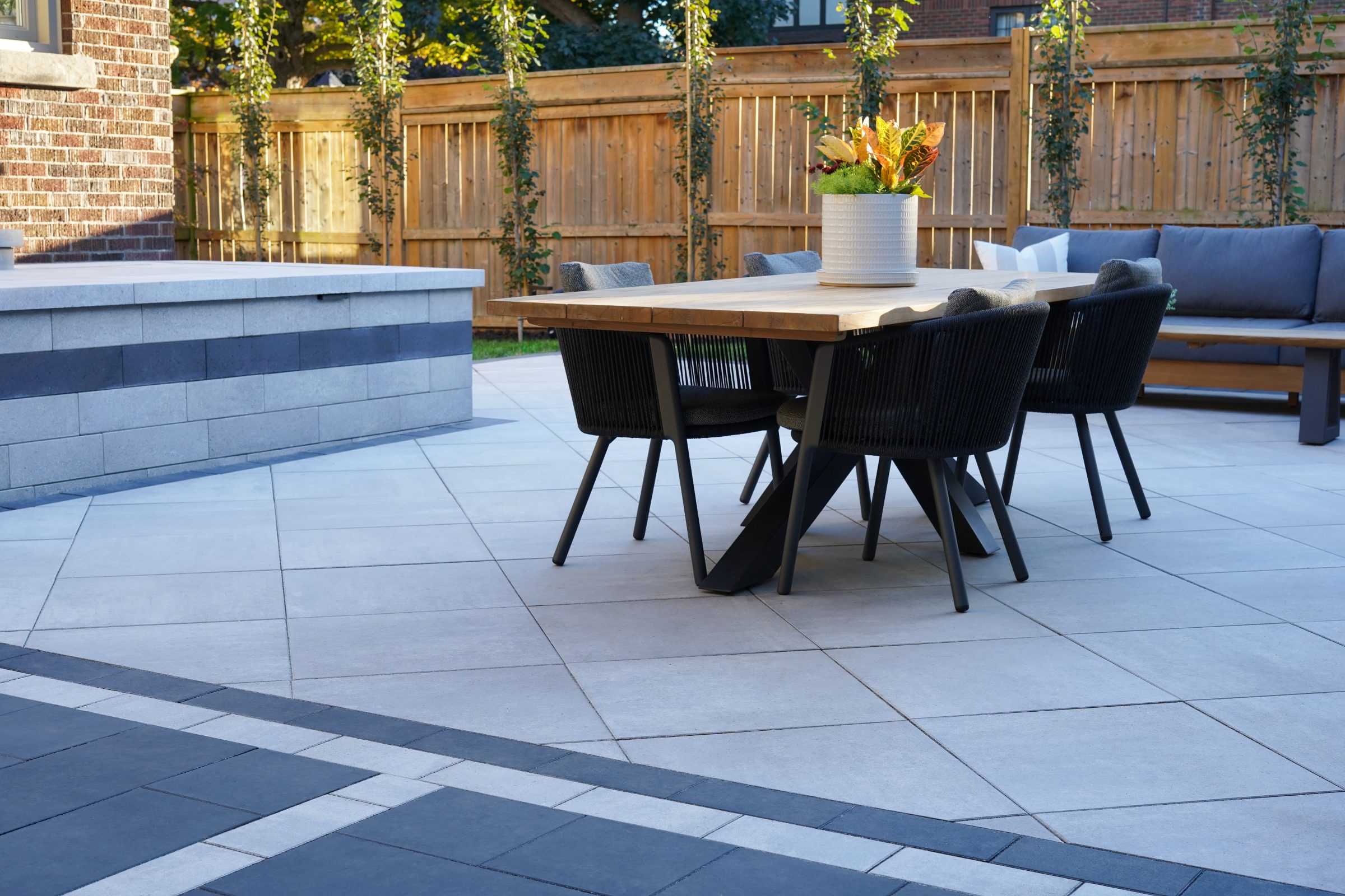 Low-angle view of a wooden dining table and black chairs on a Cloudburst patio next to a Nueva stone retaining wall.